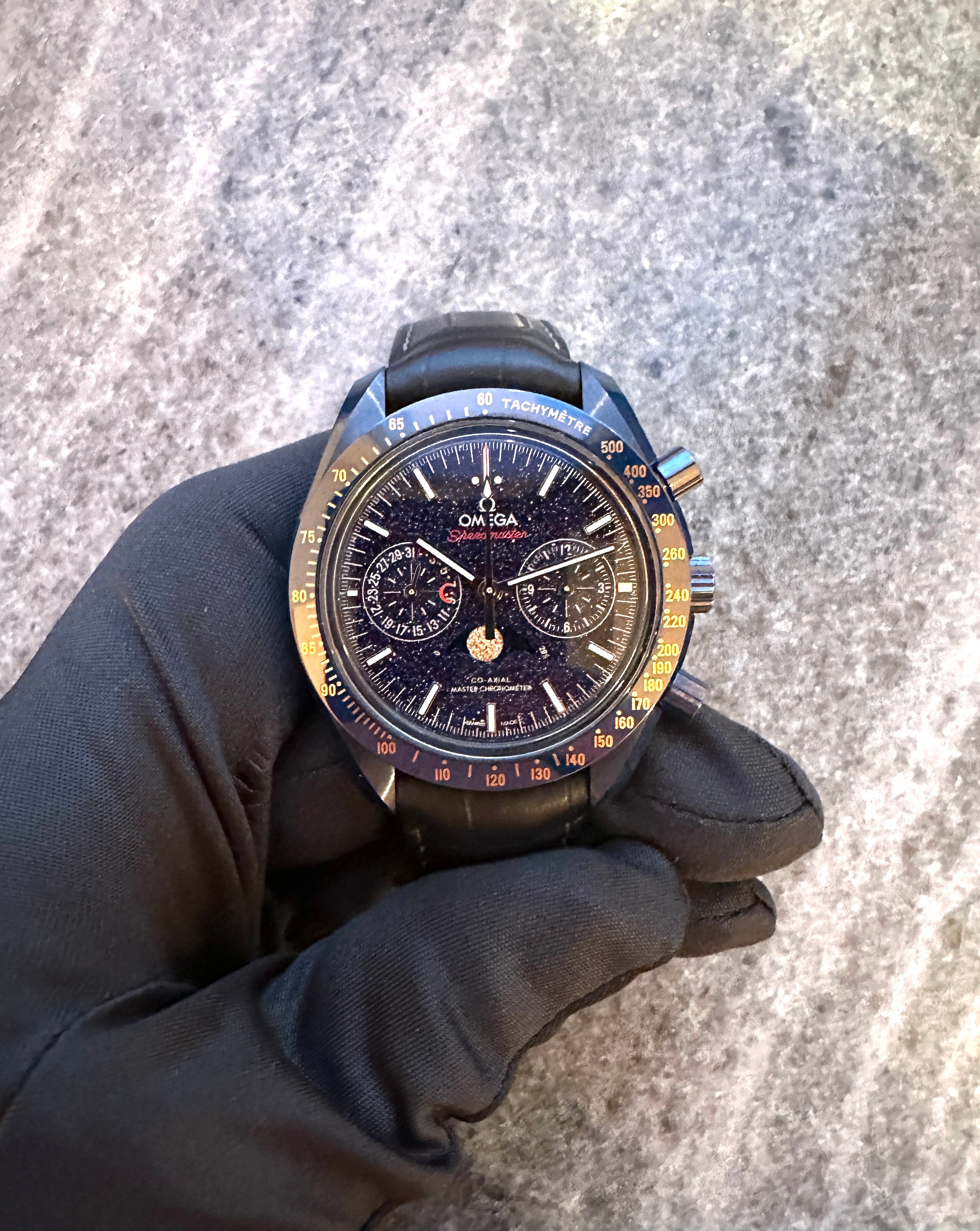 Omega Speedmaster Moonwatch Moonphase Blue Side of the Moon auf einem grauen Hintergrund, gehalten von einer Hand in einem…