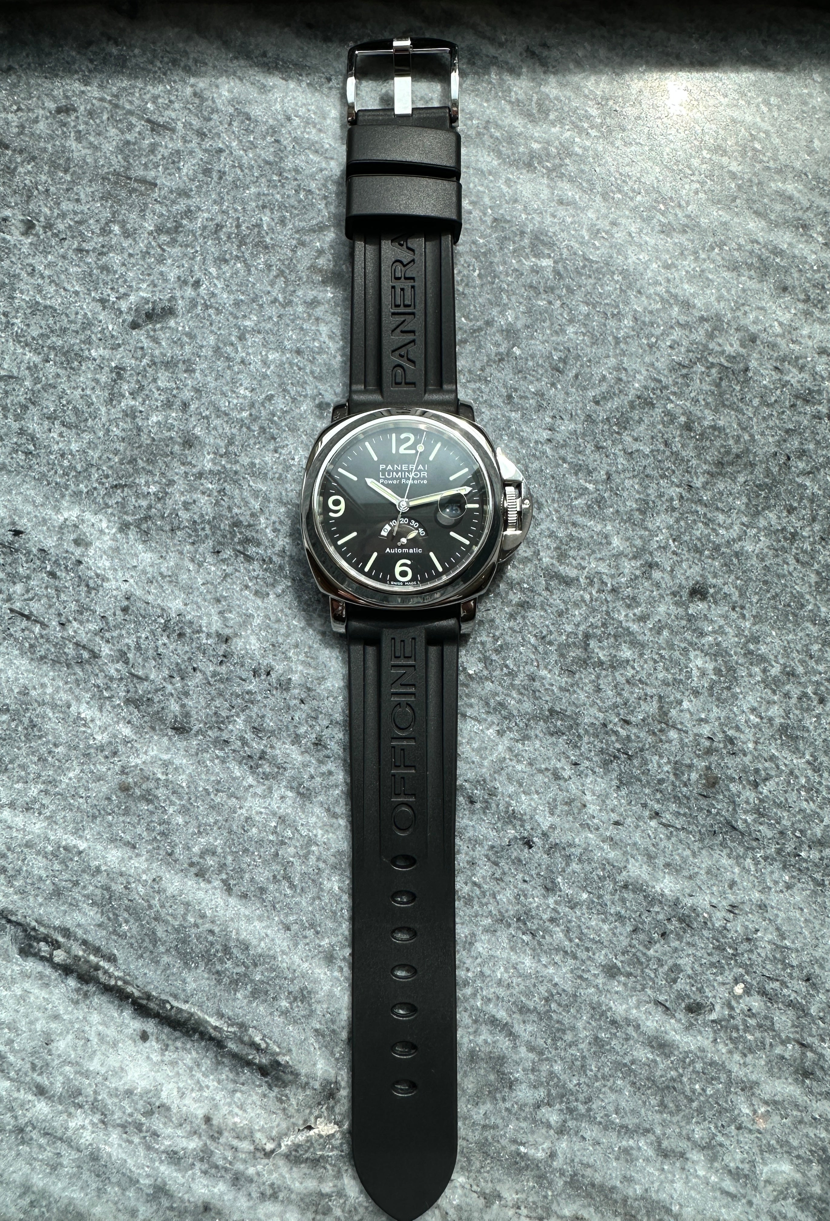 Eine Panerai Luminor Power Reserve 44mm mit schwarzem Zifferblatt und Kautschukband liegt diagonal auf grauem Steinuntergrund
