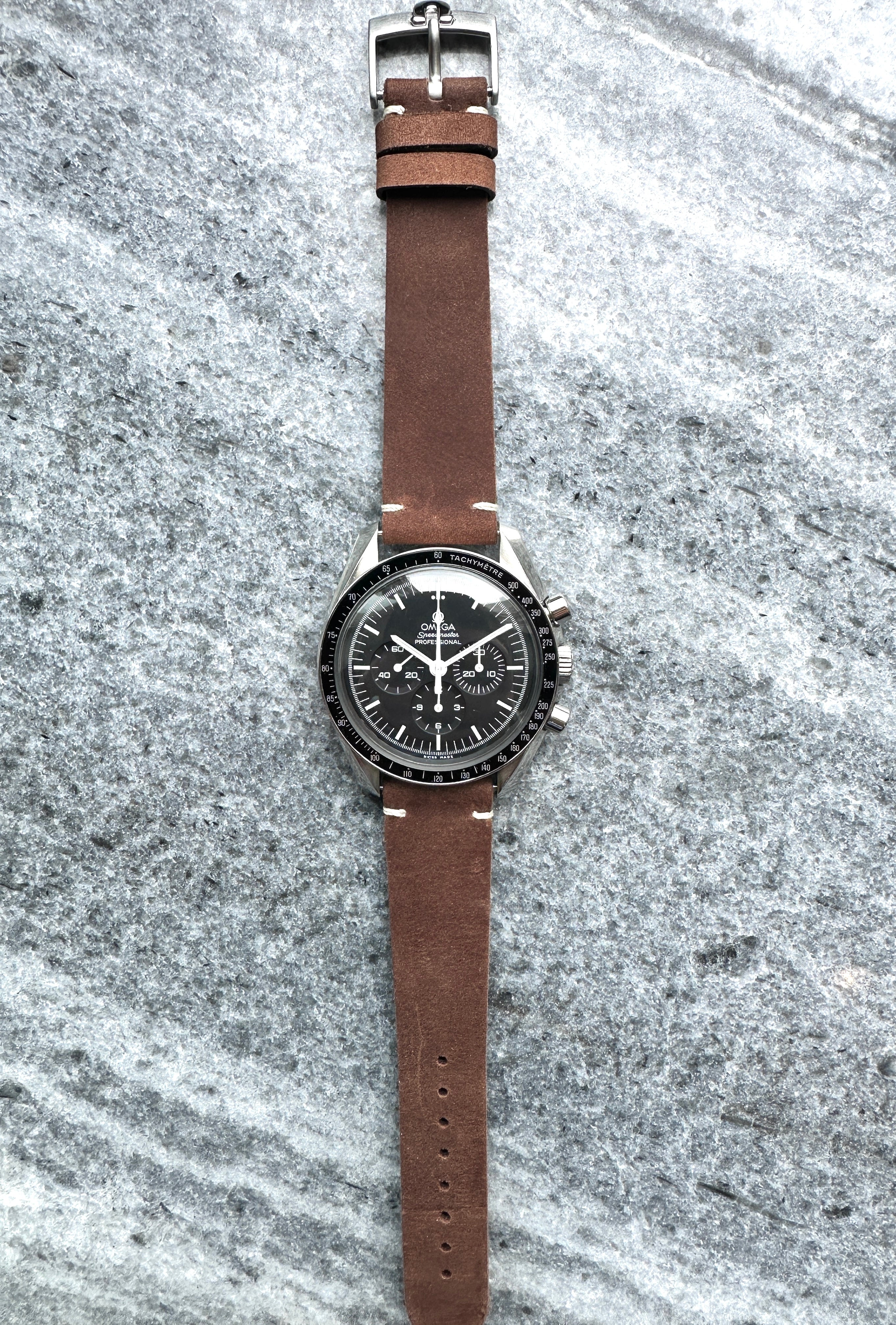 Omega Speedmaster Professional Moonwatch mit braunem Lederarmband und schwarzem Zifferblatt auf steinigem Hintergrund