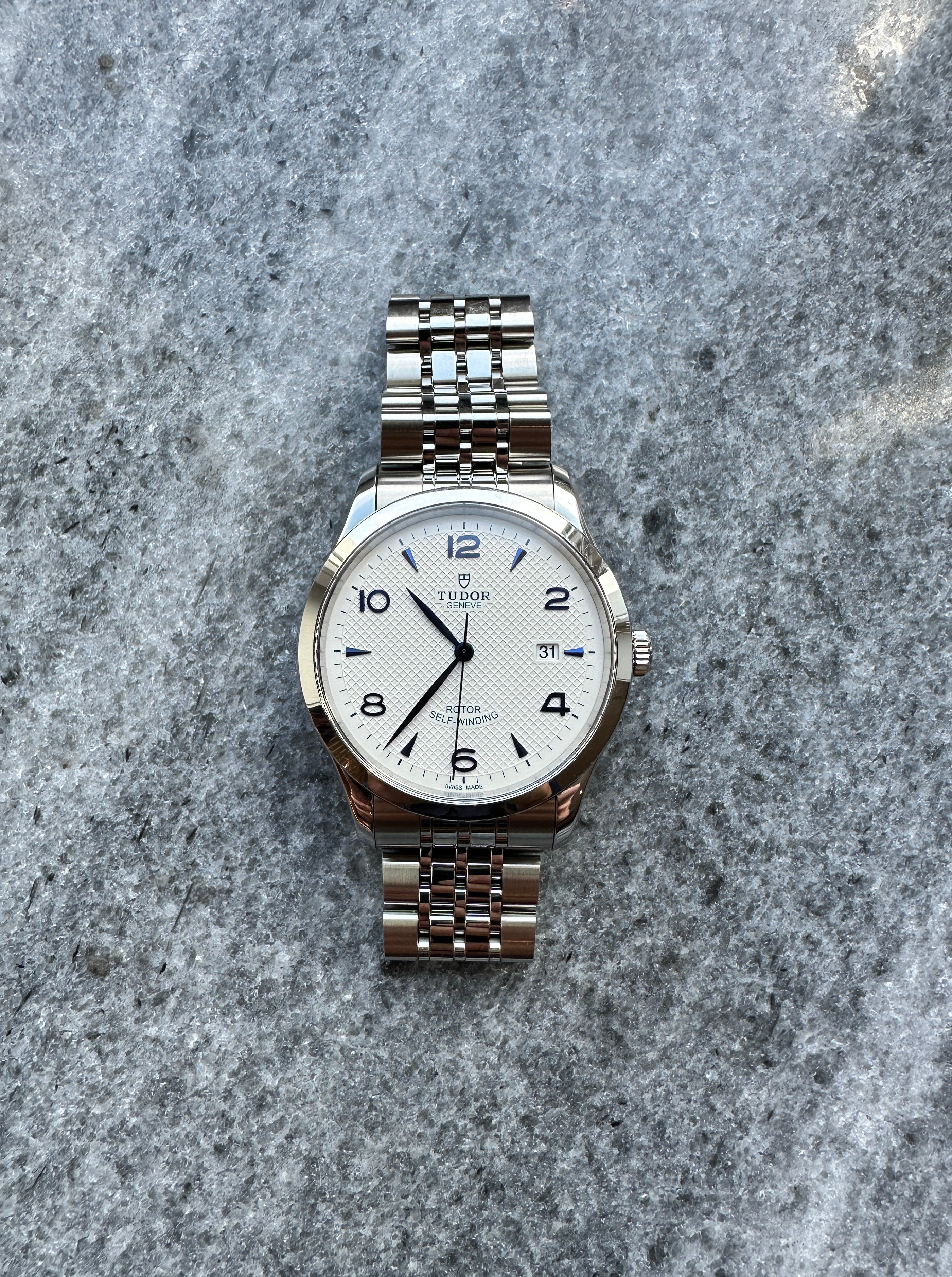 Tudor 1926 41mm Automatik Damenuhr aus Edelstahl mit weißem Zifferblatt und Jubilé-Armband auf strukturiertem grauen Hintergrund