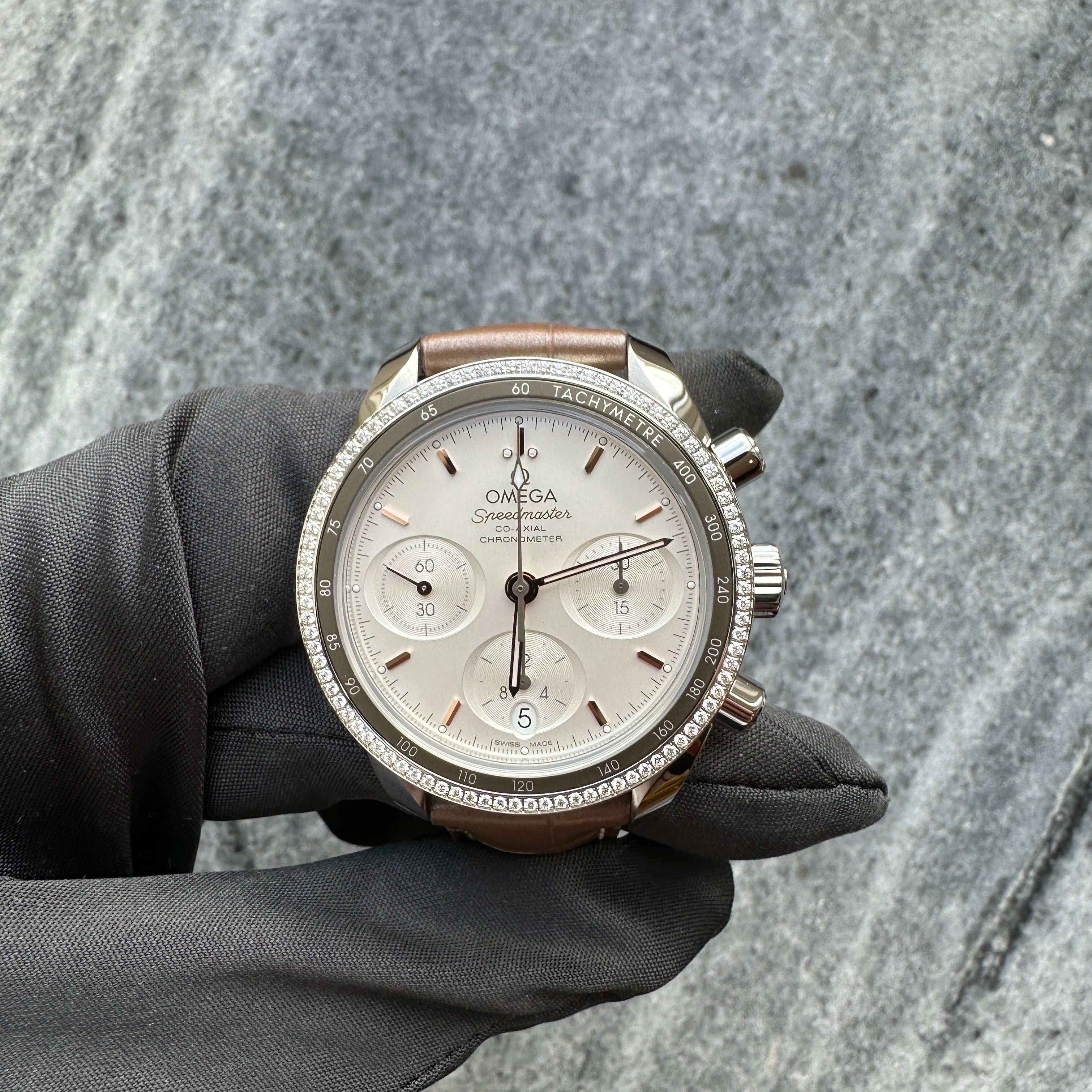 Omega Speedmaster Lady 38 Co-axial Chronograph aus Stahl mit Diamantlünette und braunem Lederarmband auf grauem Steinboden