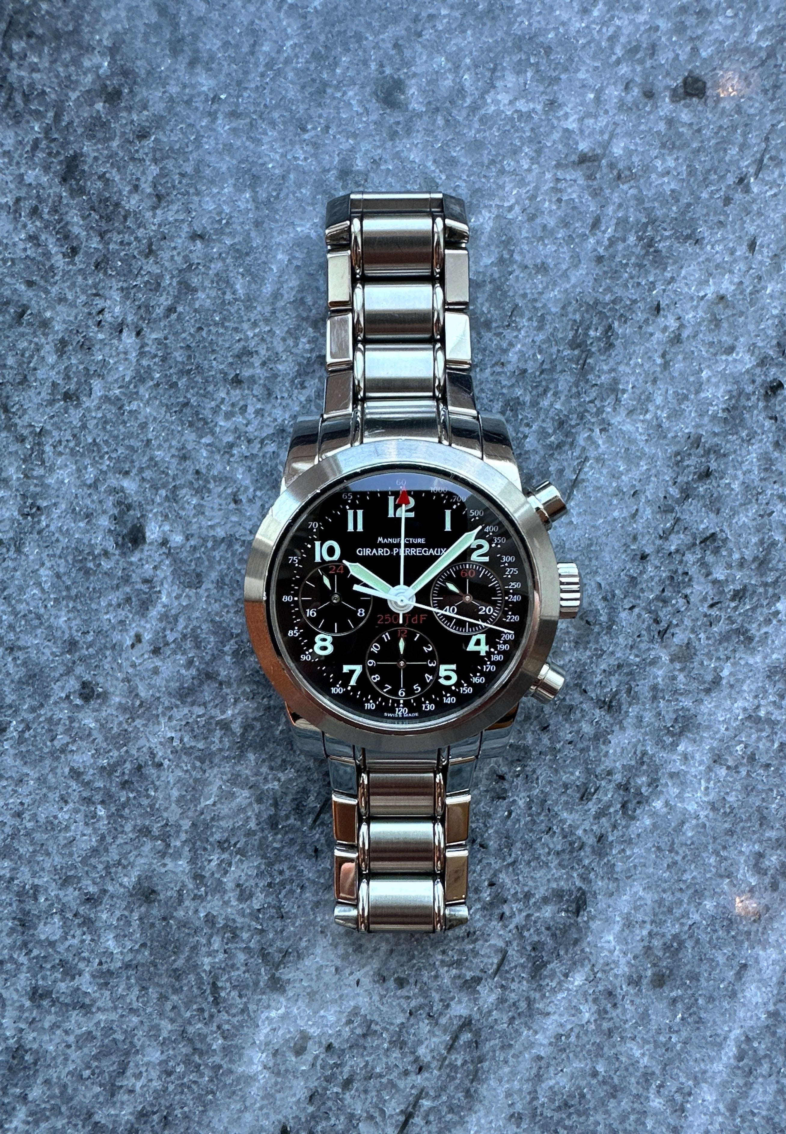 Girard Perregaux Ferrari 250, Edelstahl-Chronograph mit schwarzem Zifferblatt und Metallarmband auf grauem Stein