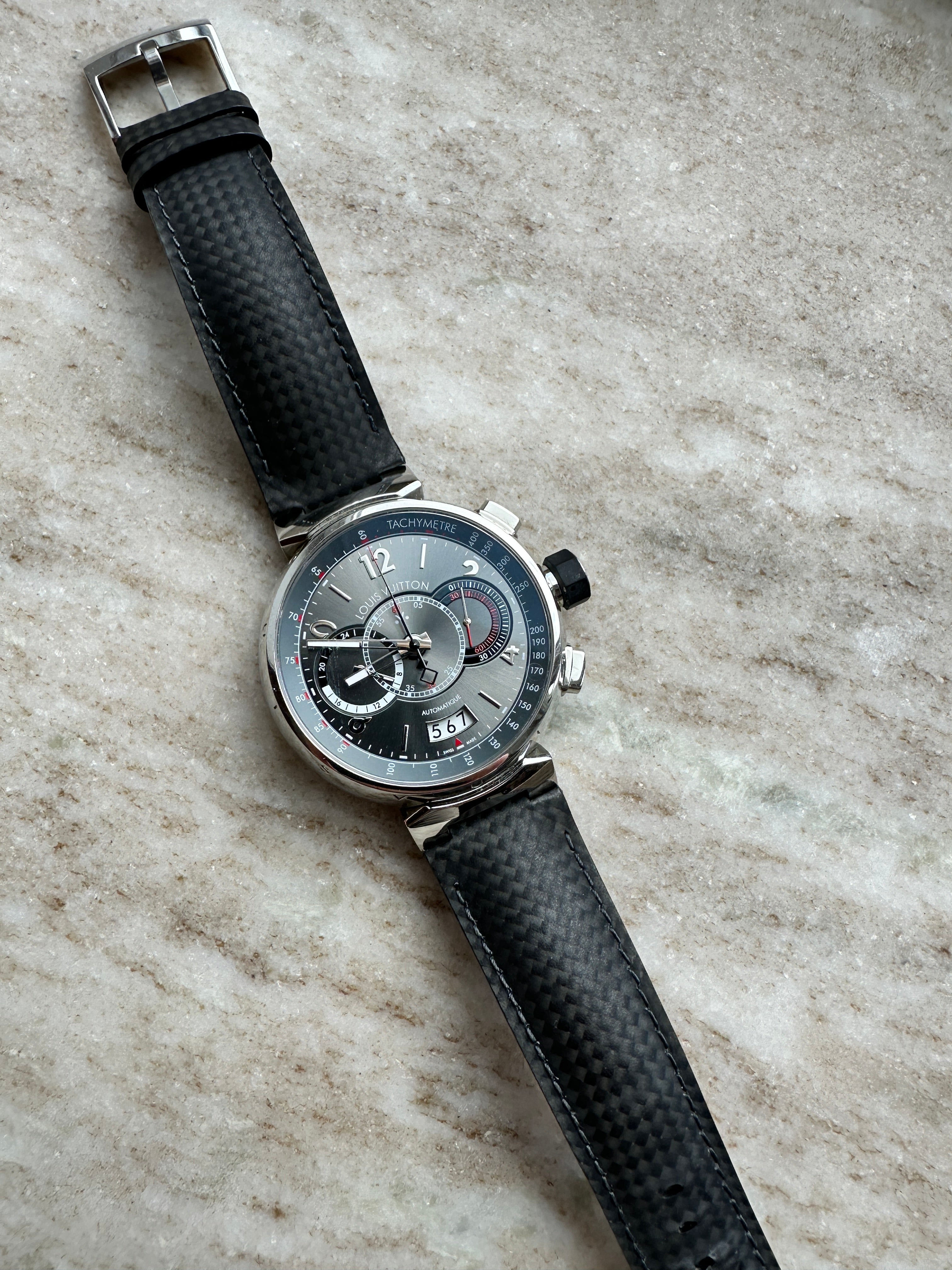 Eine Louis Vuitton Tambour Chronograph Uhr mit Edelstahlgehäuse und schwarzem Carbon-Armband liegt auf einer Marmoroberfläche