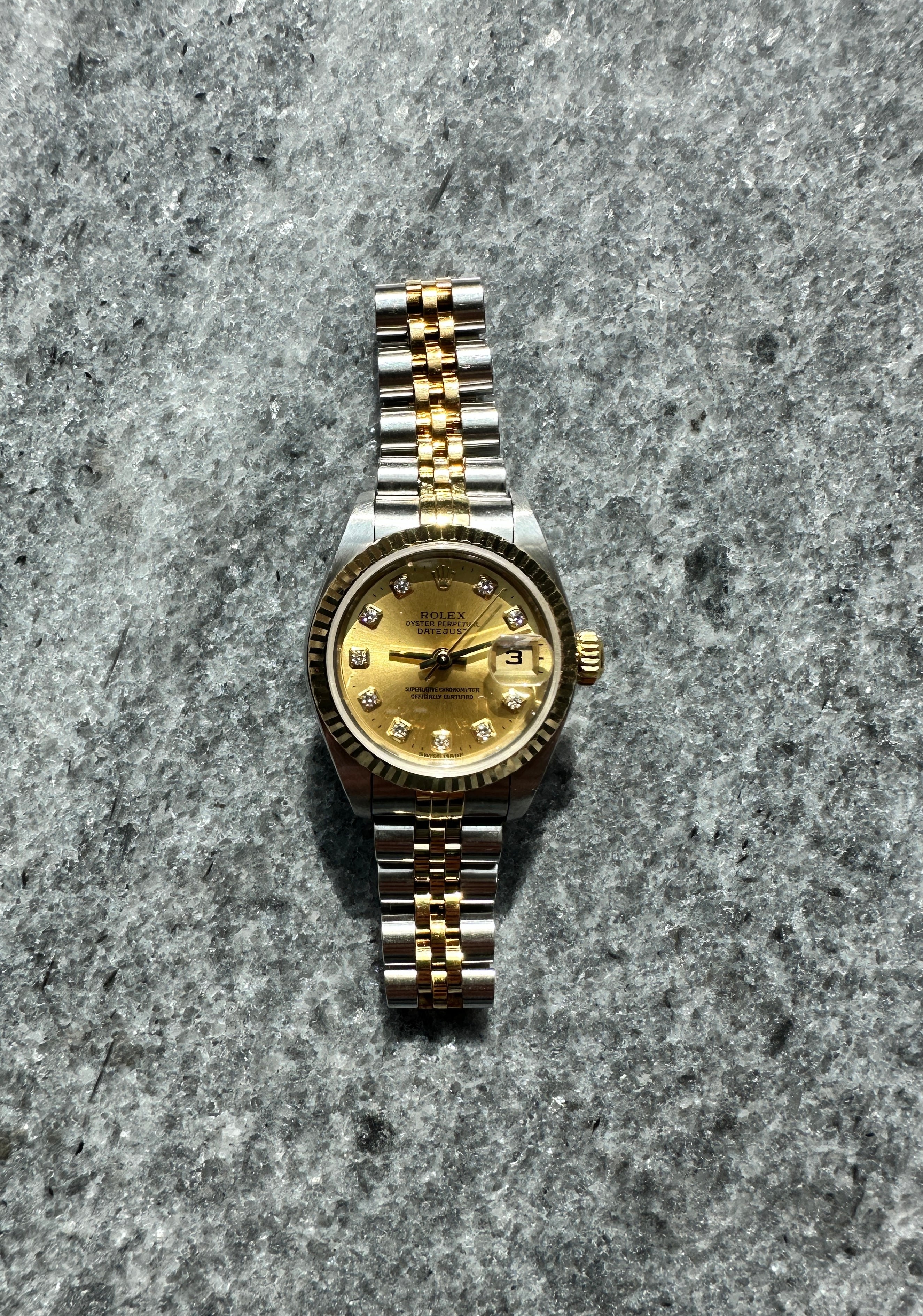 Eine Rolex Lady-Datejust Bicolor mit 10 Diamanten auf einem goldfarbenen Zifferblatt auf grauem Steinuntergrund