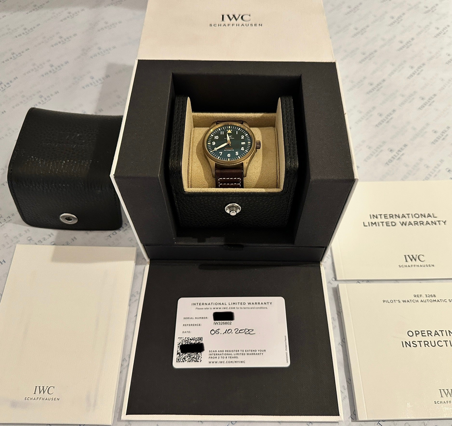 IWC Spitfire Automatic Green Bronze in geöffneter Box mit Garantiekarte und Bedienungsanleitung auf hellem Hintergrund