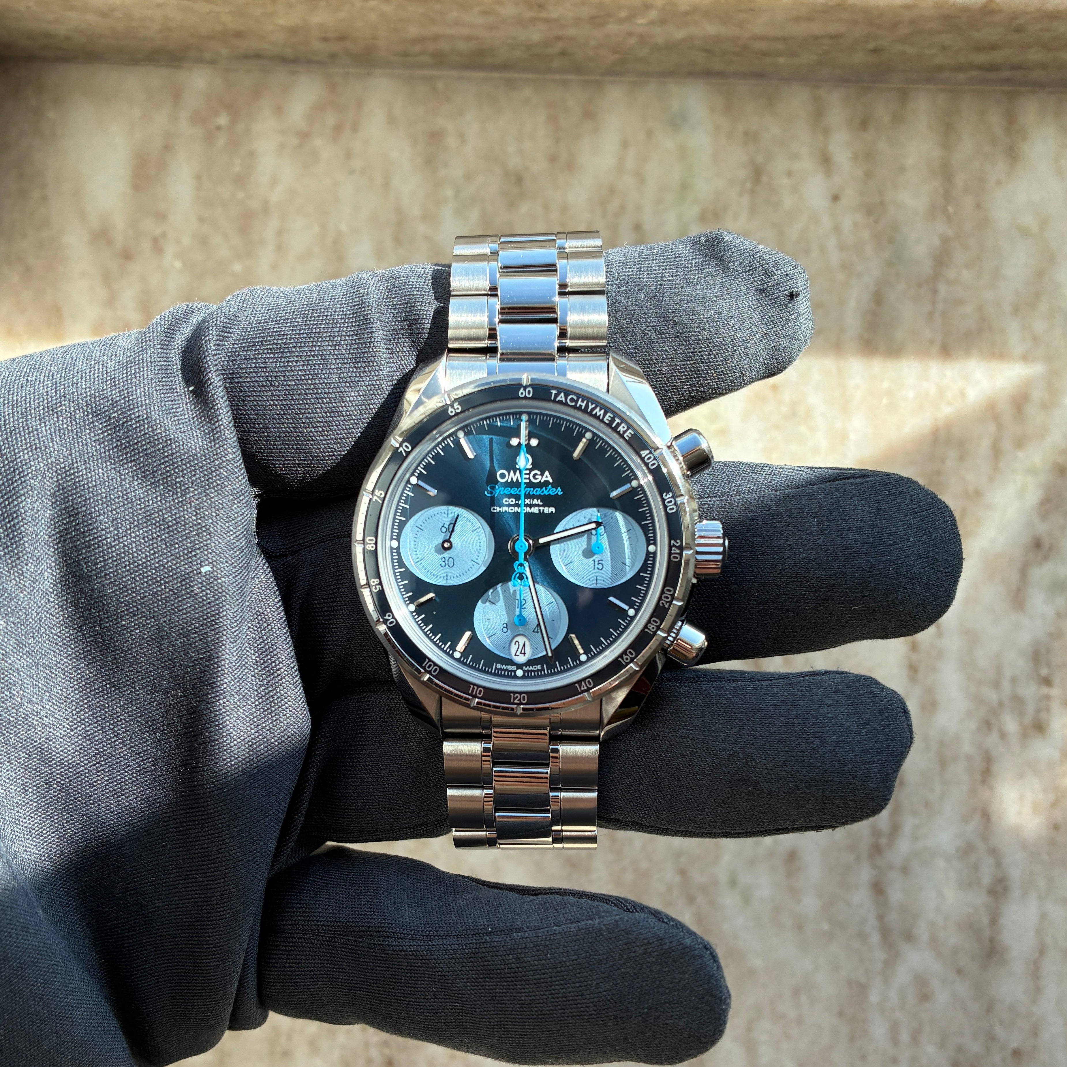 Eine Omega Speedmaster Co-Axial Automatik-Uhr mit blauem Zifferblatt und Edelstahlarmband wird von einer Hand mit schwarzer Stoffhandschuh getragen