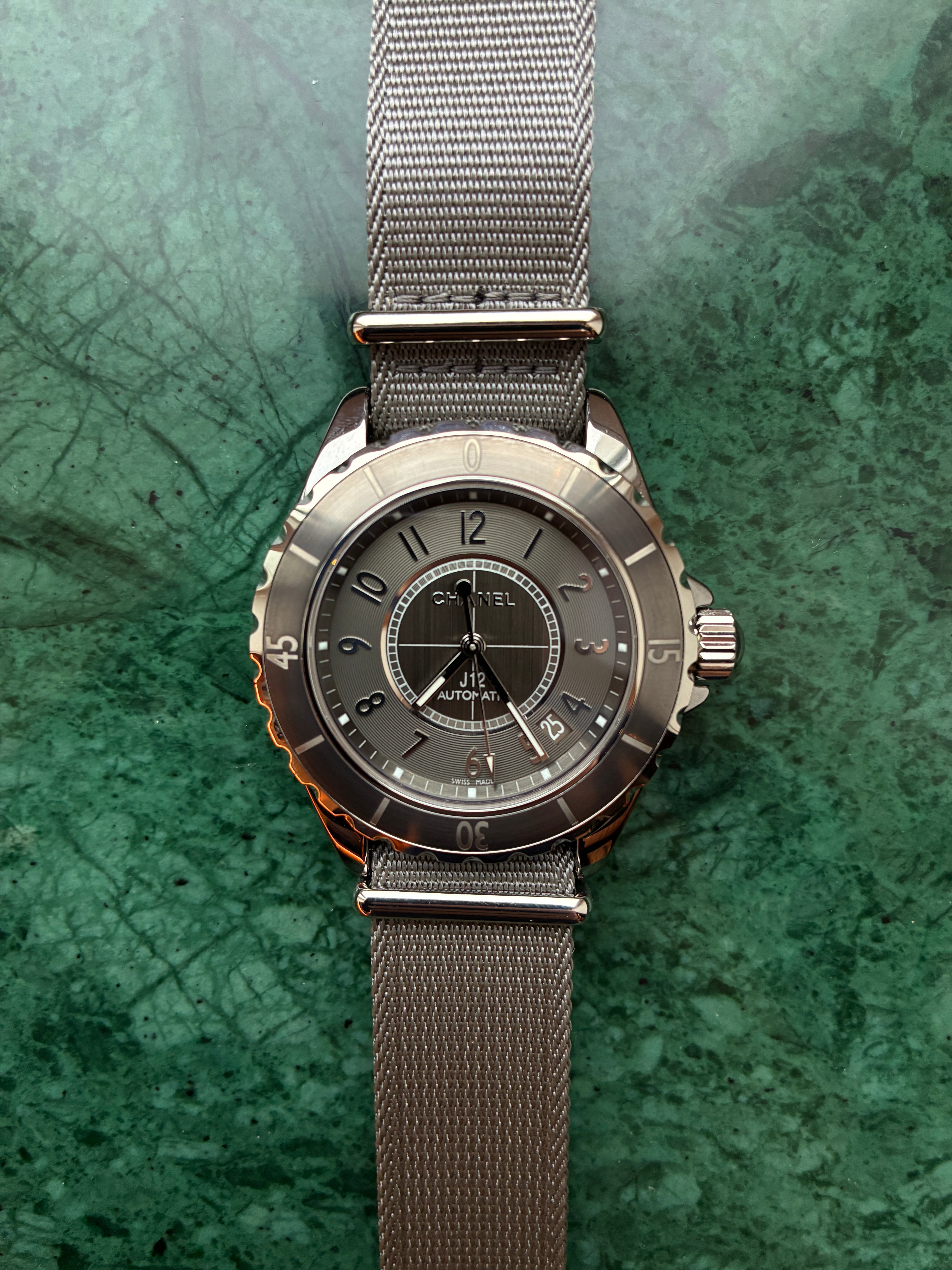 Chanel J12-g10 Uhr im Frontalansicht auf grün marmoriertem Hintergrund mit grauem Stoffarmband