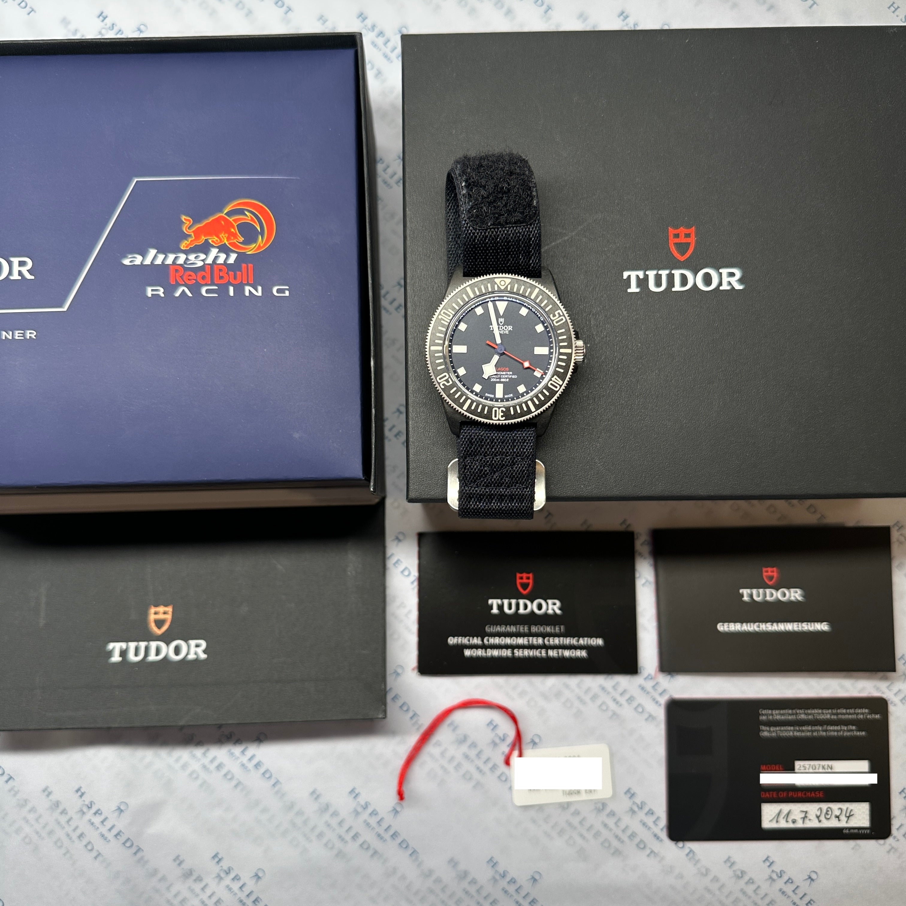 Tudor Pelagos FXD auf blauem Karton neben geöffneter Box mit Papieren und Uhr.