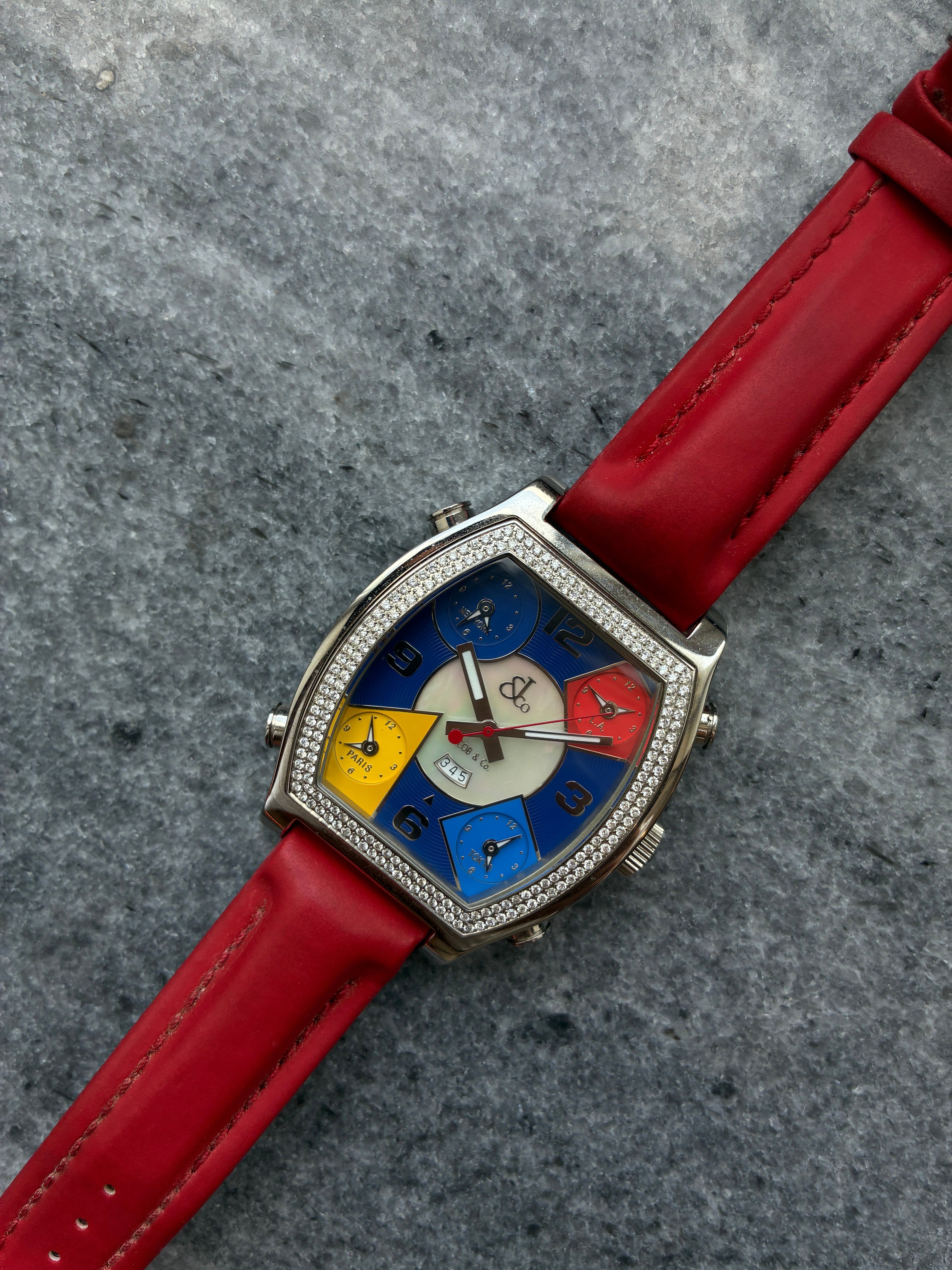 Jacob & Co. Five Time Zone Tonneau Uhr aus Edelstahl mit buntem Zifferblatt und rotem Lederarmband auf grauem Stein