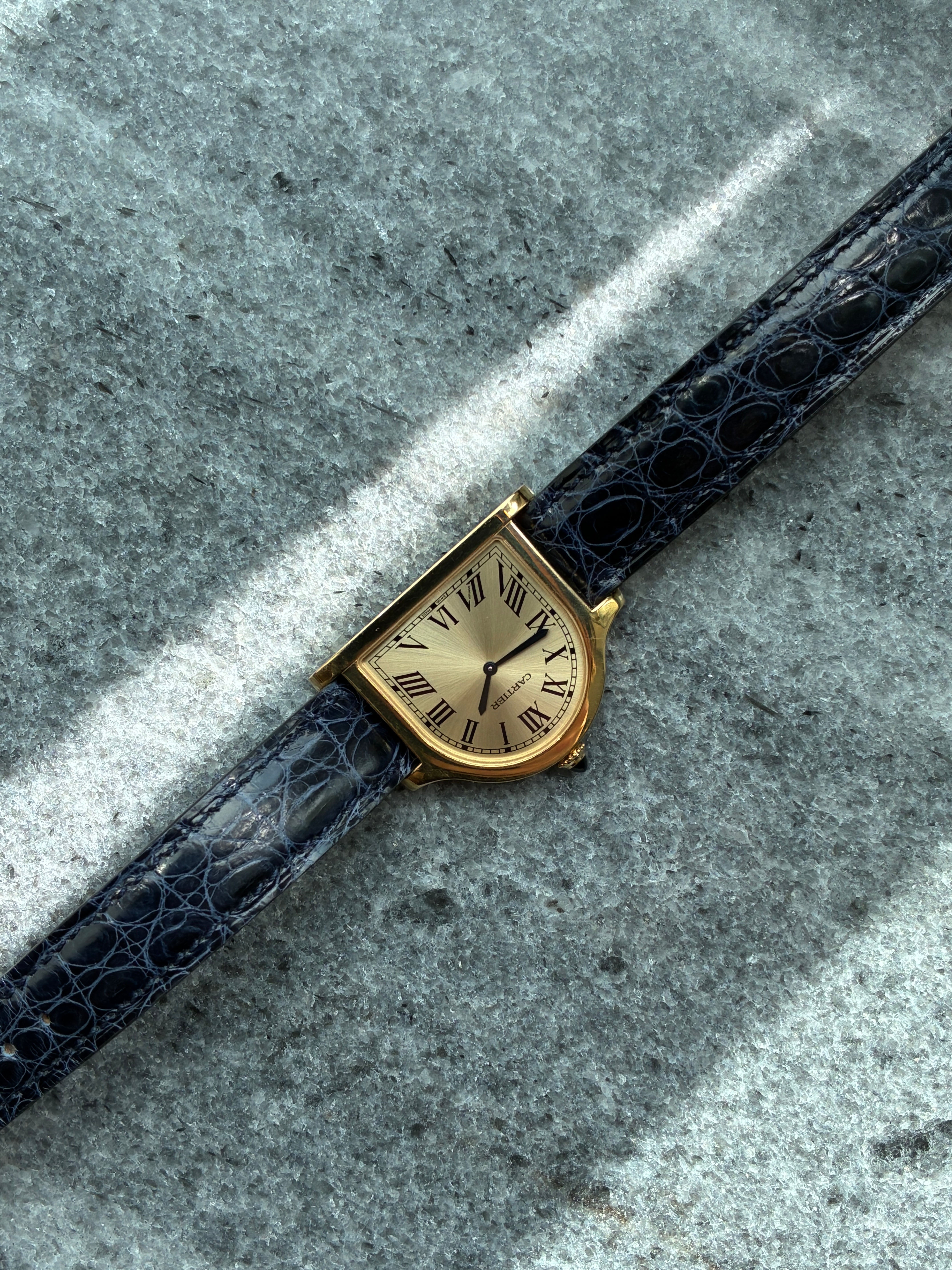 Cartier Cloche de Cartier aus Gelbgold mit blauem Kroko-Lederarmband auf grauem Stein-Untergrund