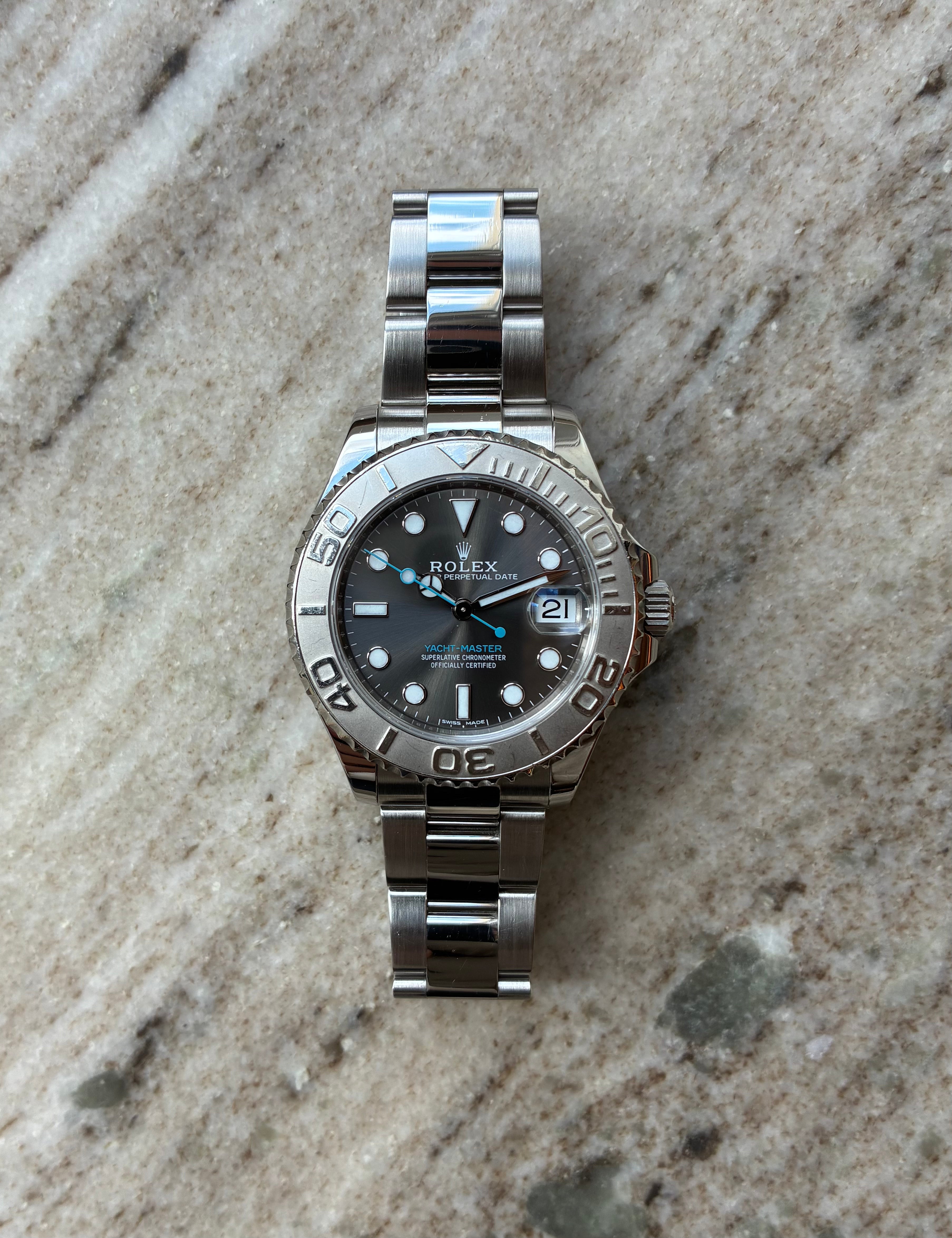 Rolex Yacht-Master 37 aus Edelstahl mit dunklem Zifferblatt und silbernem Armband auf marmoriertem Hintergrund