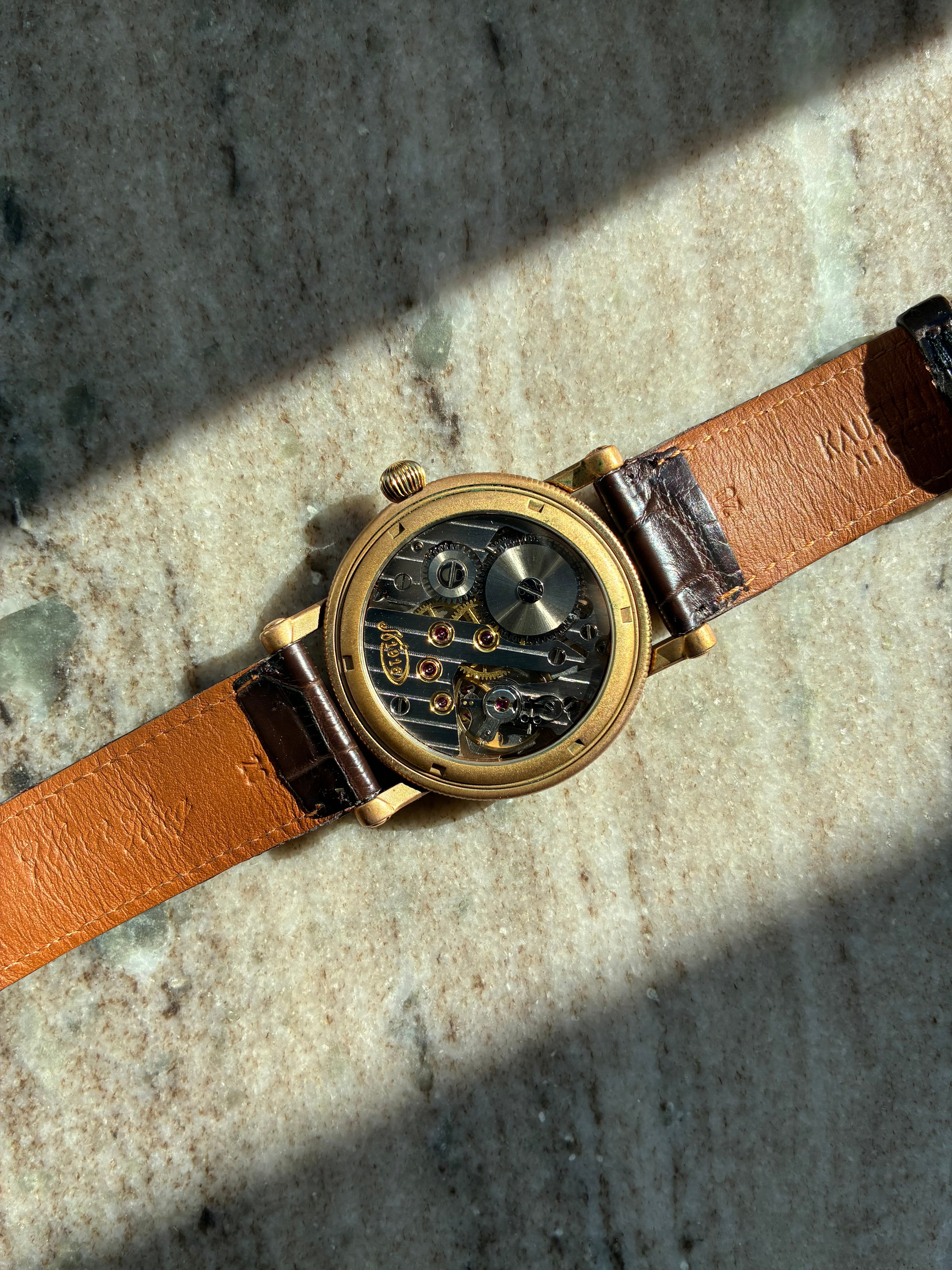 Choswiss Regulateur Bronze Rückansicht mit Sichtboden und braunem Lederarmband auf Marmorgrund
