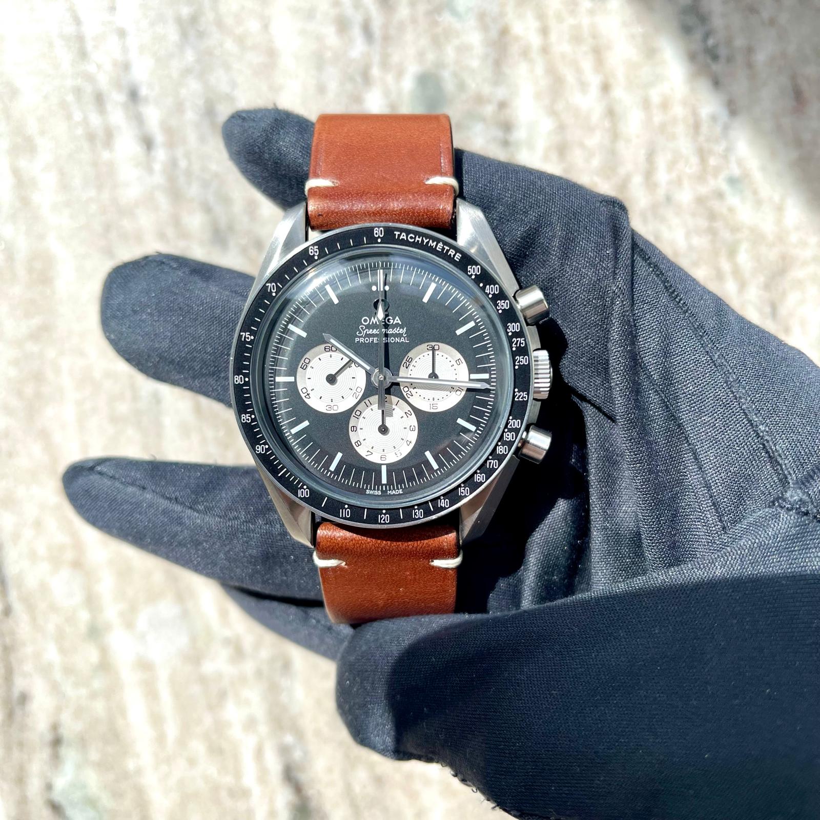 Eine Omega Speedmaster Professional Uhr mit braunem Lederarmband wird von einer schwarzen Handschuhhand gehalten, Detailansicht des Zifferblatts un…