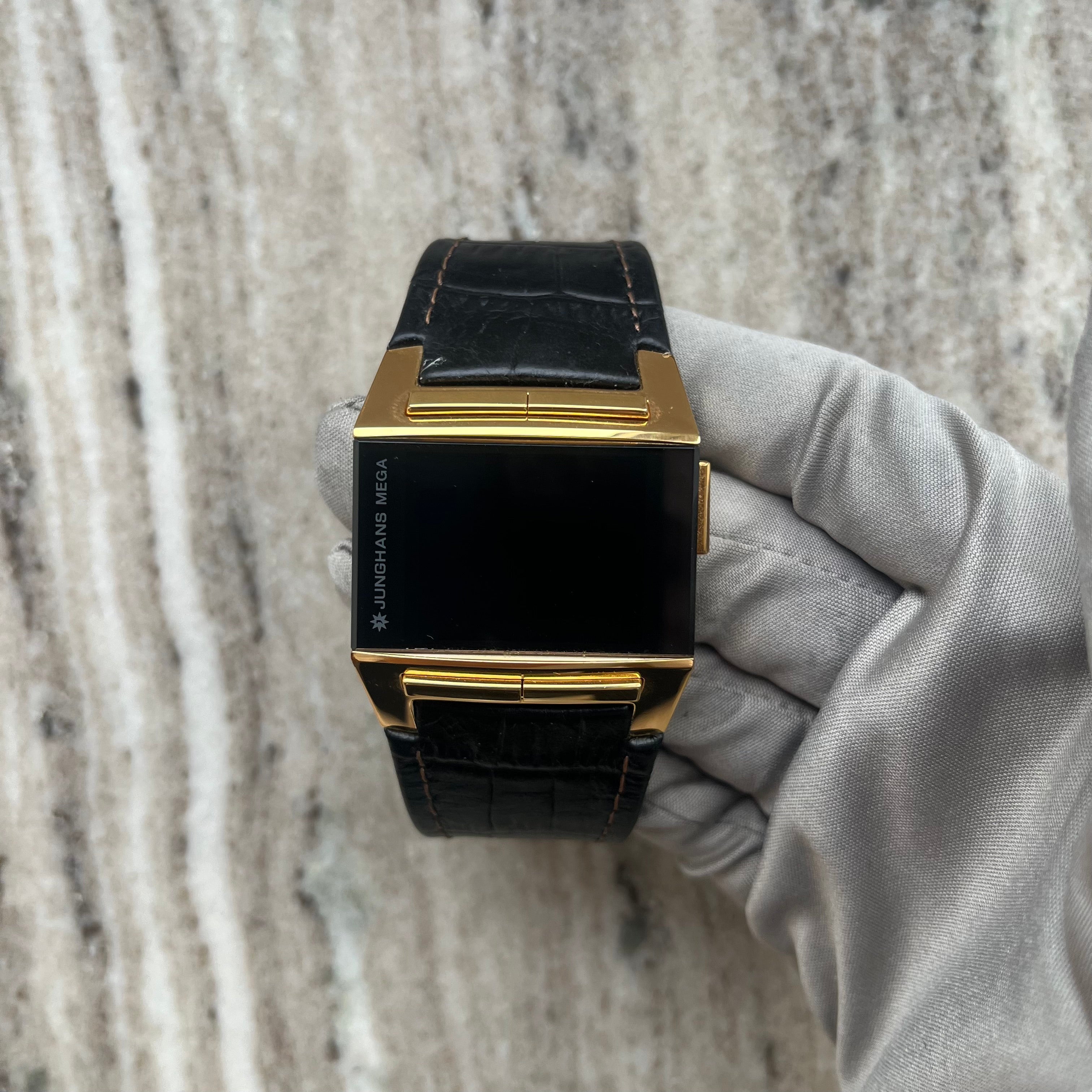 Junghans Mega 1000 Funkuhr mit schwarzem Zifferblatt und goldenem Gehäuse, gehalten in einer grauen Handschuhhand