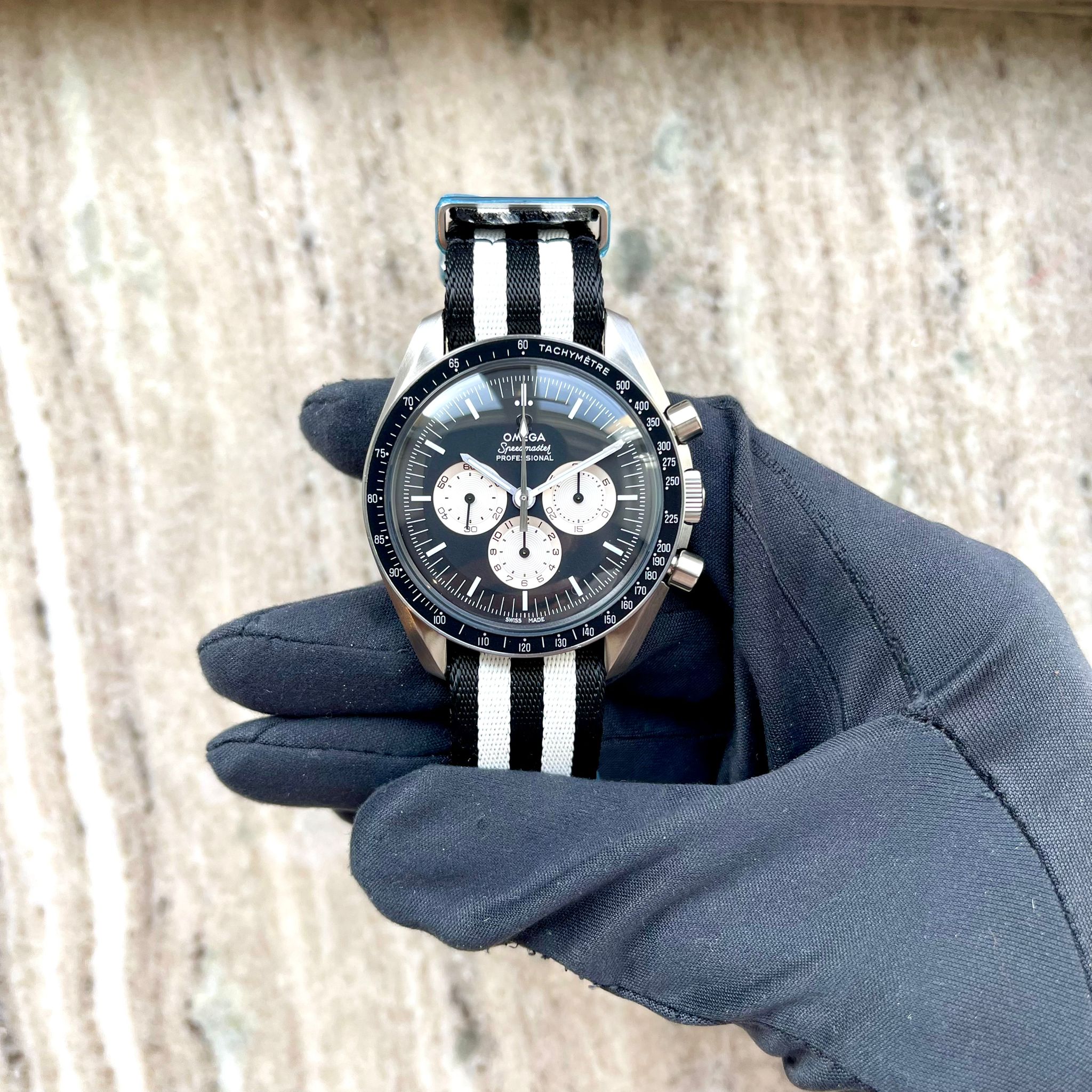 Omega Speedmaster Professional aus Edelstahl mit schwarzem Zifferblatt und bicolor-NATO-Armband, von einer Hand mit schwarzer Stoffhandschuh gehalten